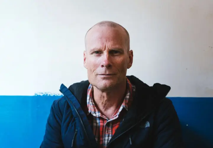 Retrato de Alex Lowe, un hombre calvo con camisa de cuadros y chaqueta azul, mirando fijamente a la cámara.