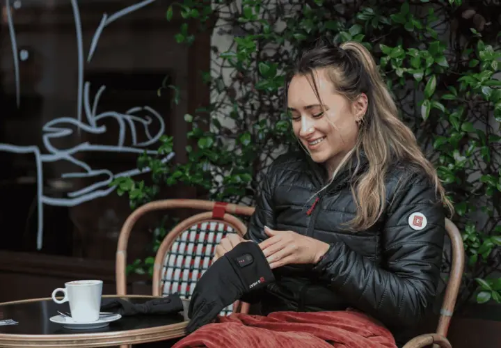 Mujer sonríe mientras se pone guantes térmicos, perfectos para el frío. Una taza de café la espera.