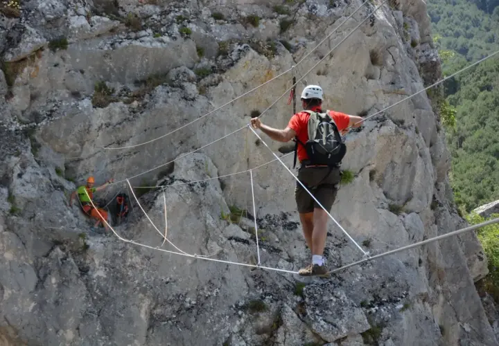 Un hombre con casco y mochila cruza un puente de cables en una pared rocosa. Otro excursionista, con casco naranja, se ve en la distancia. Esto es una demostración de que es rapel.
