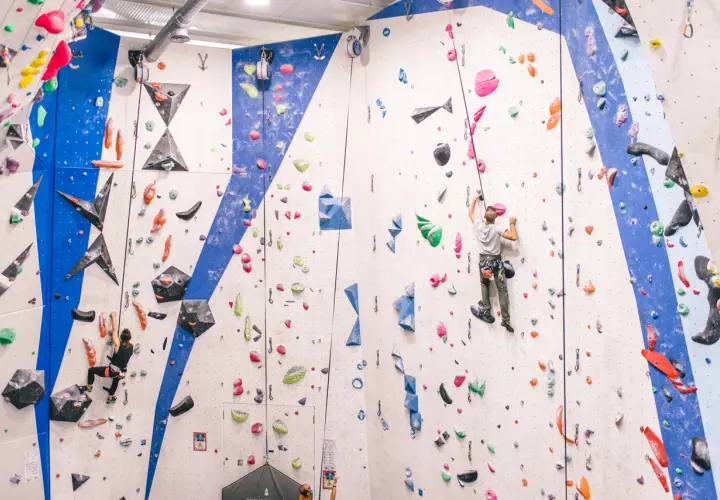 Escaladores en rocódromo, uno asciende con un asegurador automático de escalada, el otro en la pared azul.