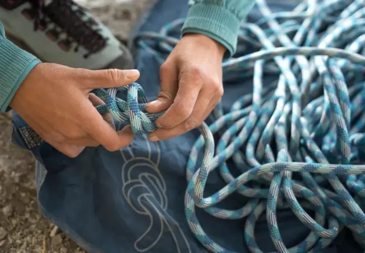 Manos preparan cuerda para machard escalada. Botas de montaña y lona azul con nudos dibujados.