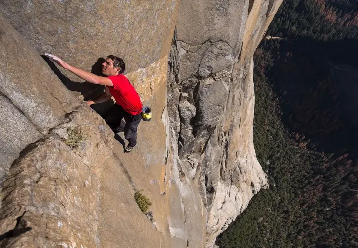 Un escalador en solitario asciende una pared de roca imponente, capturando la esencia de un documental de escalada. La vista es vertiginosa, con un bosque denso abajo.