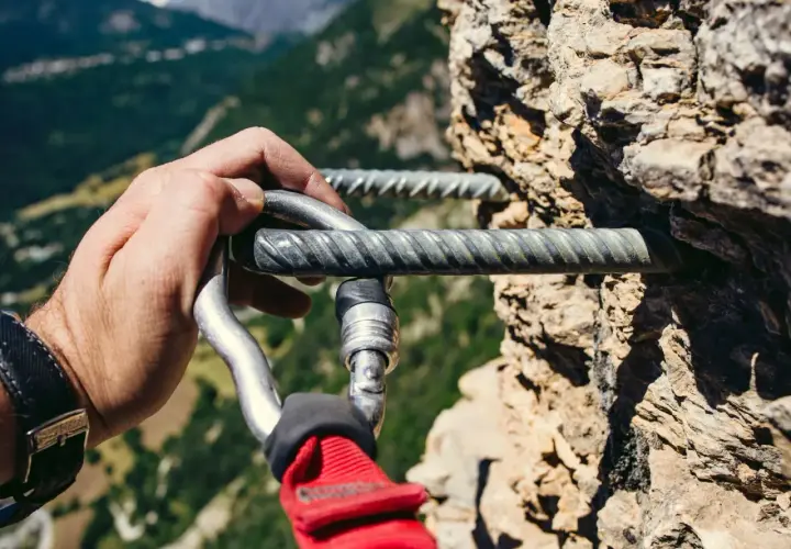 Mano enguantada se engancha a la **línea de vida vía ferrata** en una pared rocosa, con un paisaje montañoso al fondo.