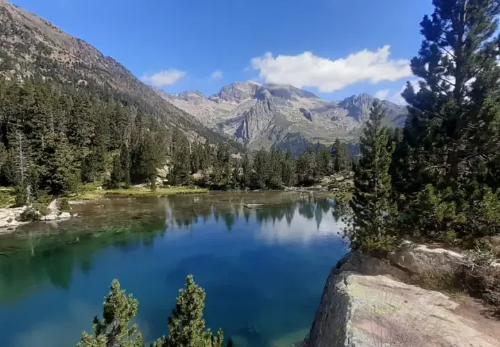 El ibonet de Batisielles, un espejo de agua turquesa rodeado de pinos y montañas imponentes, refleja el cielo azul y las nubes.