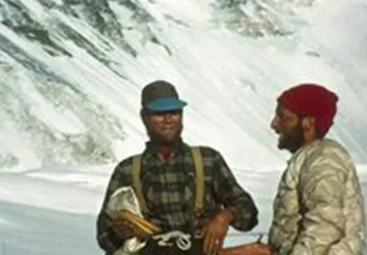 Dos montañistas sonríen en la nieve. Uno, con gorra y camisa de cuadros, sostiene un pico. El otro, con gorro rojo, parece ser Tom Hornbein.