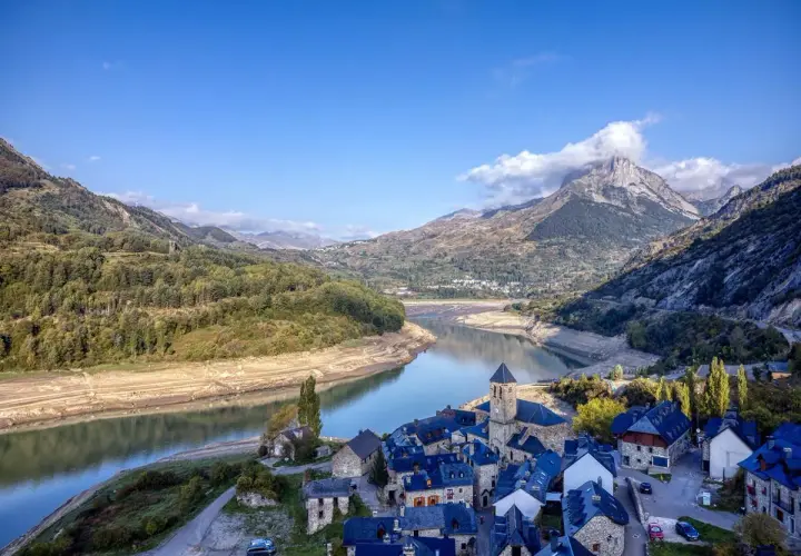 Pueblo de piedra junto a un embalse, con montañas nevadas de los picos de Huesca al fondo.