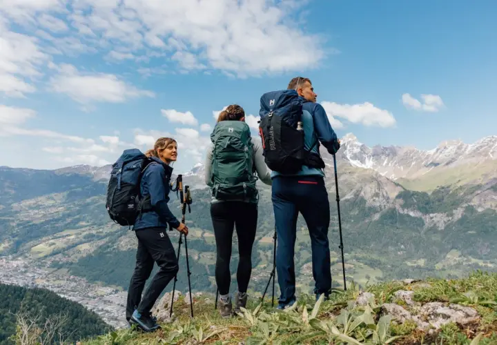 Tres excursionistas con mochilas y bastones admiran un paisaje montañoso. Sus prendas, ideales para distintos tipos de goretex, los protegen del clima.