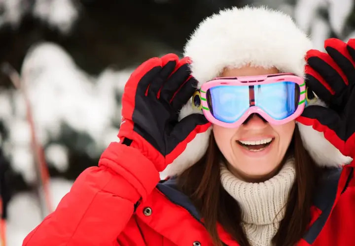 Mujer sonriente con gorro de piel y gafas de esquí rosas, lista para la nieve. Explora las categorías de gafas de esquí para encontrar tu estilo.