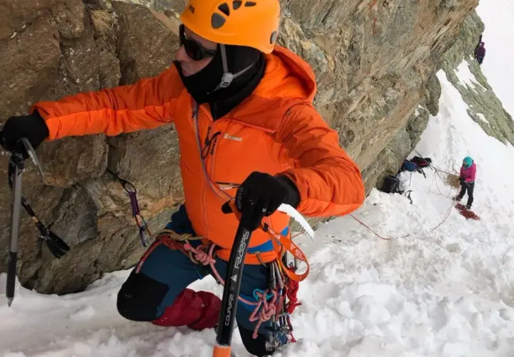 Alpinista con casco naranja y chaqueta brillante usa un piolet para escalar una pared rocosa nevada.