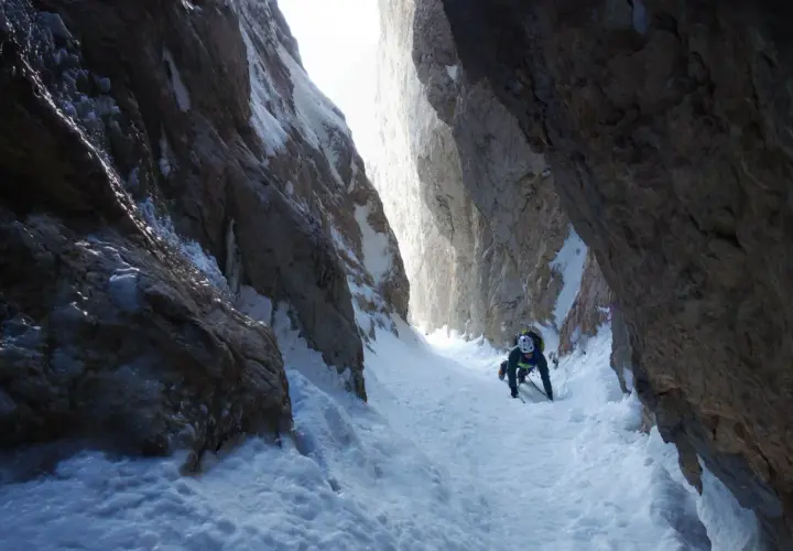 Alpinista asciende por un estrecho cañón nevado, usando piolet y crampones. La estaca de nieve se ve firme bajo sus pies.