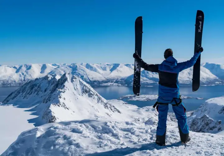Persona en la cima de una montaña nevada, sosteniendo un splitboard que es un equipo versátil para ascender y descender.