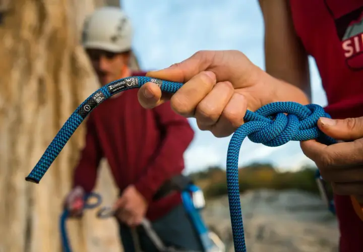Manos atando nudos de escalada con una cuerda azul. En el fondo, otro escalador con casco.