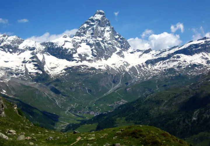 Majestuoso pico nevado se alza sobre un valle verde con un pueblo. El camino de senderismo gr5 etapes serpentea entre las colinas.