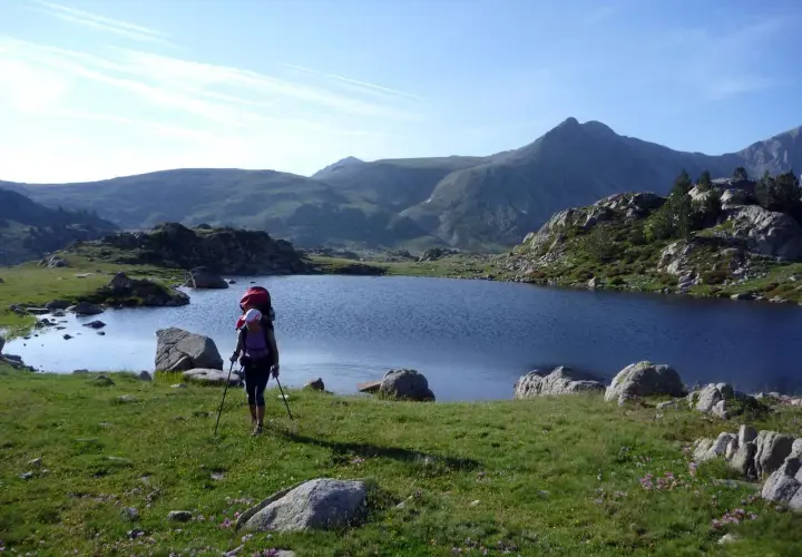 Senderista con mochila y bastones camina por un prado verde hacia un lago azul en los Pirineos de GRP Andorra.
