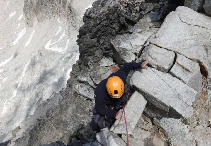Alpinista con casco naranja escala roca, buscando el aneto por coronas.