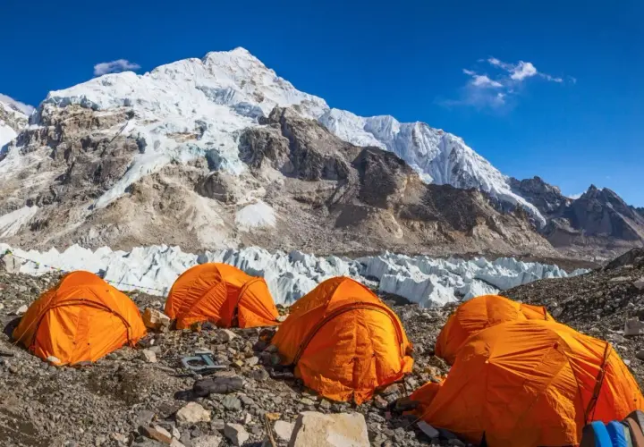 Campamento de expedición en la base de la cordillera más alta del mundo, con tiendas naranjas y glaciares imponentes bajo un cielo azul.