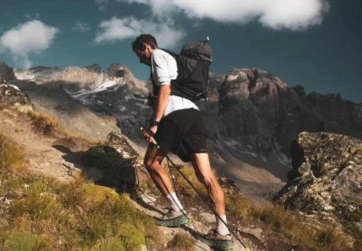 Kilian Jornet asciende una ladera rocosa en los Pirineos, con bastones y mochila, bajo un cielo azul con nubes.