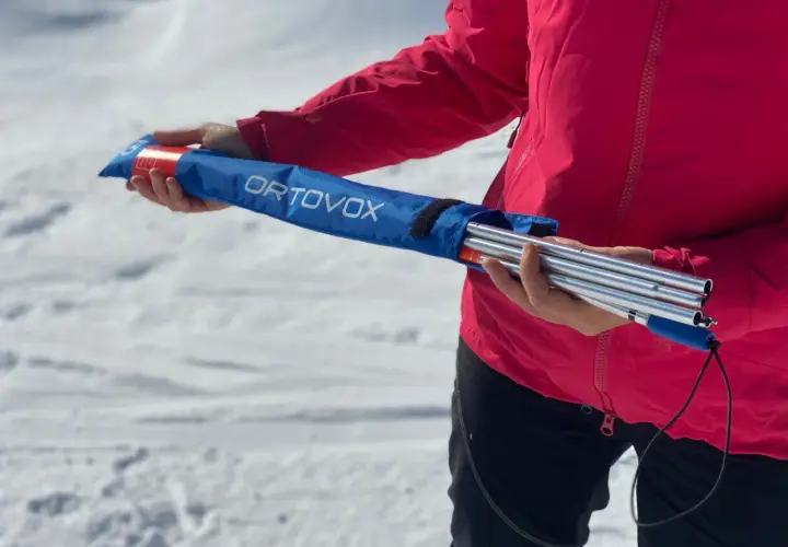 Persona con chaqueta rosa sostiene una arva, sonda y pala ORTOVOX en una bolsa azul, lista para la aventura en la nieve.
