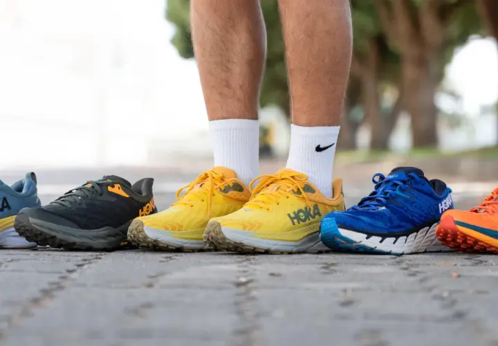 Cinco pares de zapatillas Hoka con gran amortiguación, en colores azul, gris, amarillo, azul y naranja, alineados en un camino.