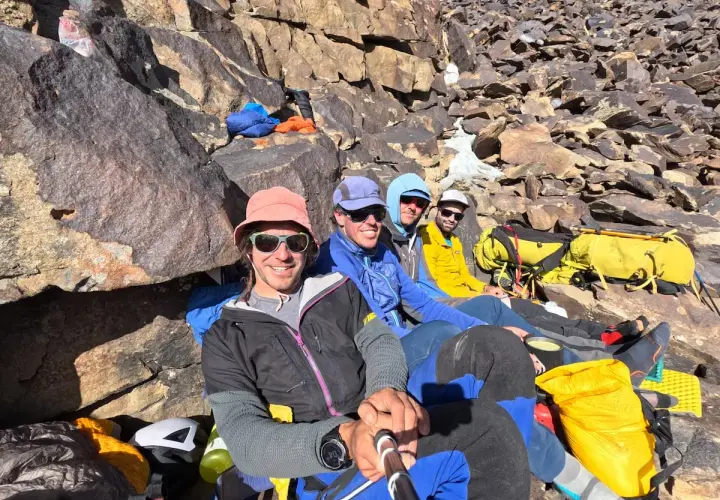 Cuatro montañistas sonríen en la ladera rocosa, listos para la aventura. El **bru busom** de uno de ellos, un sombrero rosa, destaca entre el equipo de expedición.
