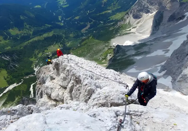 Escaladores en una vía ferrata, asegurados a un cable de acero que recorre la cresta rocosa. El paisaje montañoso es espectacular.