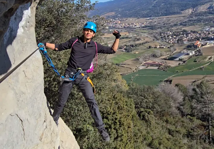 Hombre con casco azul y arnés, asegurado a un cable de **material vía ferrata**, disfruta de las vistas del valle.