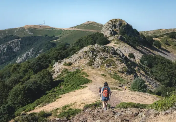 Senderismo en montaña con bastones, comparando rutas en AllTrails vs. Wikiloc. Paisaje verde y rocoso bajo un cielo azul.