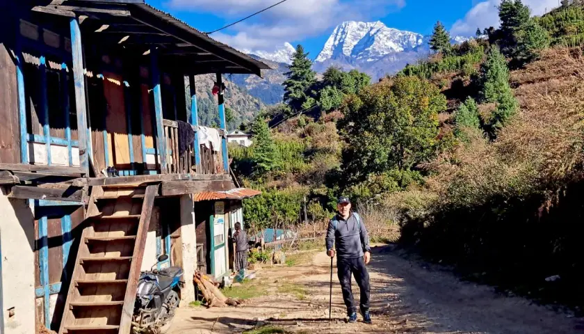 Hombre con bastón de trekking en un camino rural, con una casa tradicional y montañas nevadas al fondo. Una aventura de trekking Himalaya.