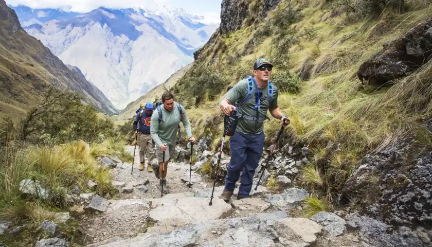Tres excursionistas suben un sendero rocoso en las montañas, usando bastones de trekking para ayudarse.