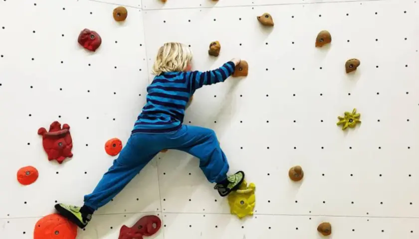 Niño rubio con camiseta a rayas azules y pantalón azul practica escalada para niños en un rocódromo blanco con presas de colores.