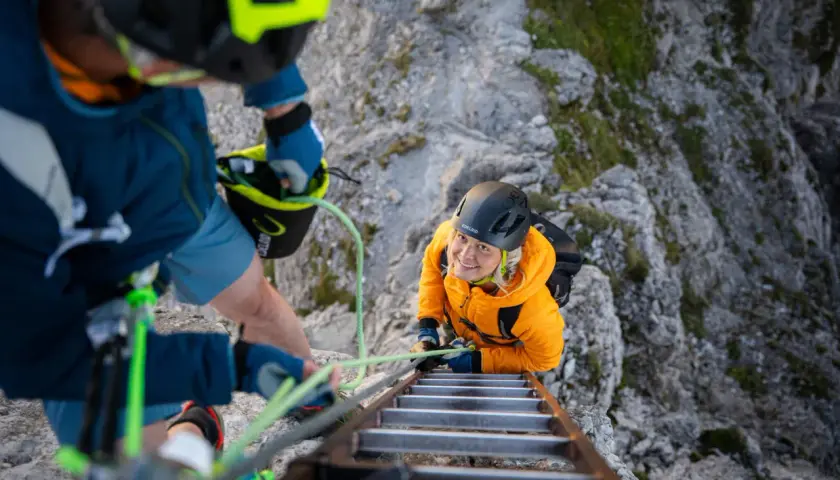 Dos personas con equipo ferrata escalan una pared rocosa, una subiendo una escalera metálica y la otra asegurándola.