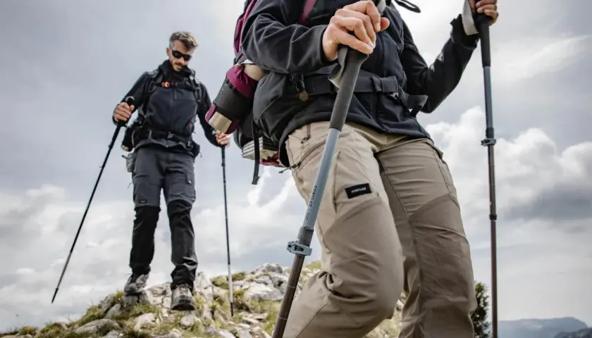 Dos excursionistas suben una ladera rocosa, usando sus pals per caminar para mayor estabilidad. El cielo nublado sugiere una aventura desafiante.