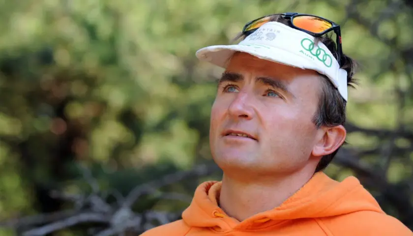 Ueli Steck, con gorra blanca y sudadera naranja, mira hacia arriba con determinación.