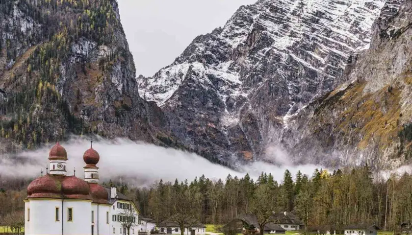 Iglesia blanca con cúpulas rojas junto a un lago, rodeada de bosques y majestuosas montañas nevadas en los Alpes alemanes.