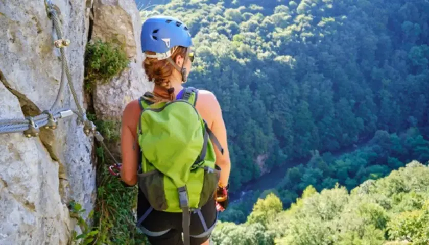 Mujer con casco y mochila verde en vía ferrata, con vistas a un bosque frondoso. Afortunadamente, no hubo ningún accidente de escalada.