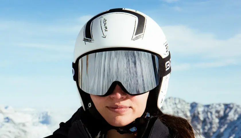 Mujer con casco de esquí blanco y gafas espejadas, lista para deslizarse por la montaña.