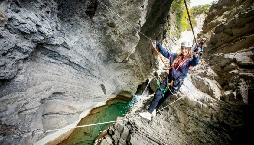 Mujer sonríe mientras cruza un barranco usando técnicas de escalada y tirolina. Otro escalador la sigue.