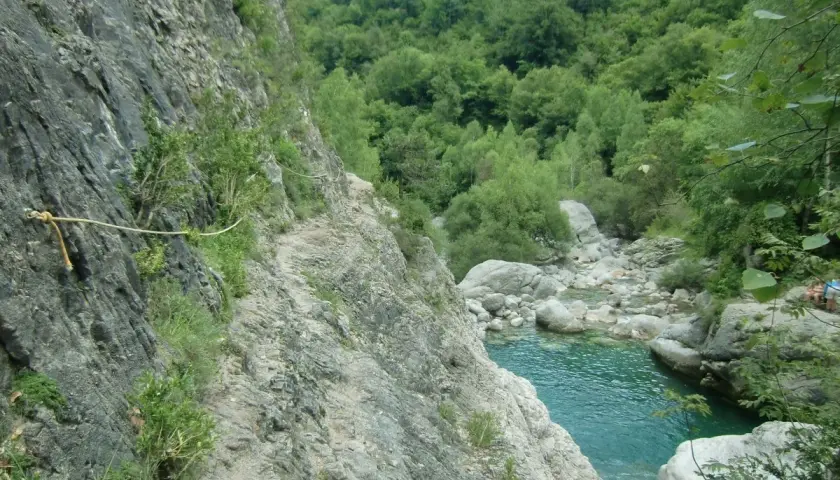 Sendero rocoso con cuerda de seguridad a lo largo de la pared de la garganta de Escuain, con un río turquesa y vegetación exuberante al fondo.