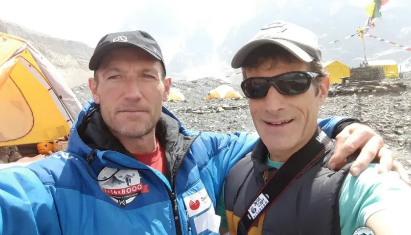 Alberto Zerain y un compañero posan sonrientes en un campamento de montaña, con tiendas amarillas y banderas de oración al fondo.