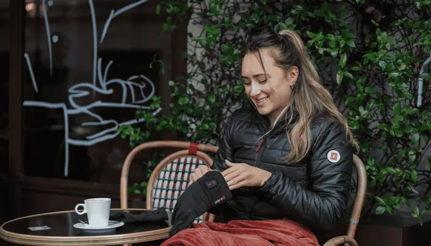 Mujer sonríe mientras se pone guantes térmicos, perfectos para el frío. Una taza de café la espera.