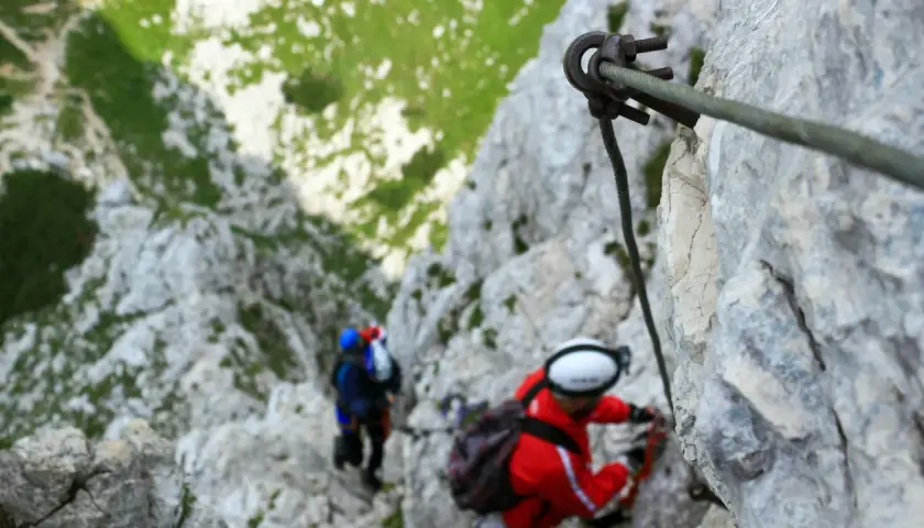 Dos personas en una vía ferrata de iniciación, aseguradas a un cable de acero mientras ascienden por una pared rocosa.