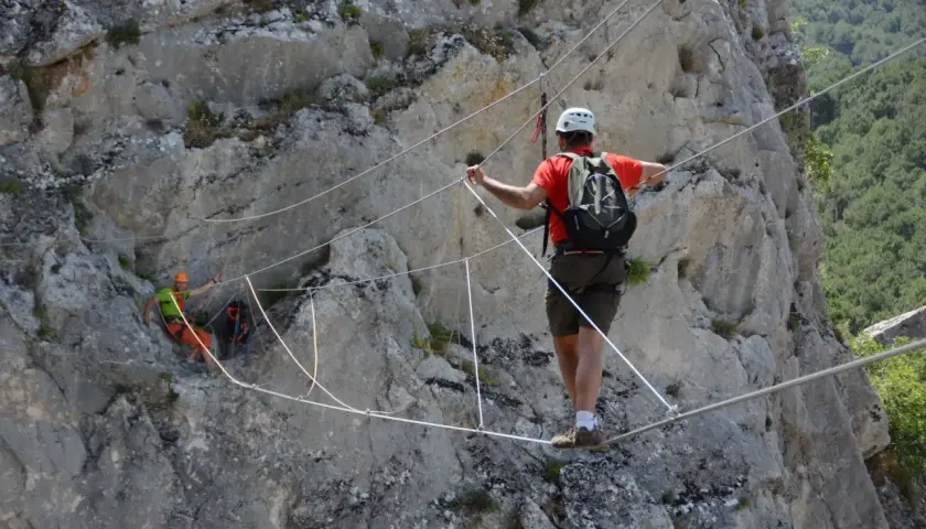Un hombre con casco y mochila cruza un puente de cables en una pared rocosa. Otro excursionista, con casco naranja, se ve en la distancia. Esto es una demostración de que es rapel.