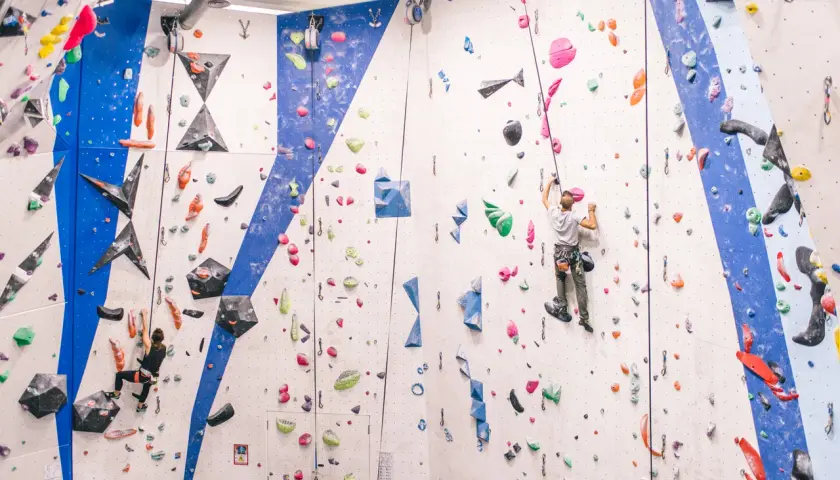 Escaladores en rocódromo, uno asciende con un asegurador automático de escalada, el otro en la pared azul.