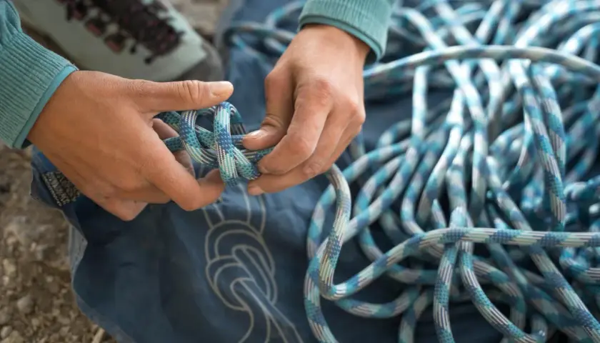 Manos preparan cuerda para machard escalada. Botas de montaña y lona azul con nudos dibujados.