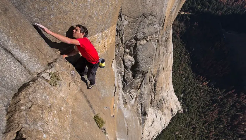 Un escalador en solitario asciende una pared de roca imponente, capturando la esencia de un documental de escalada. La vista es vertiginosa, con un bosque denso abajo.
