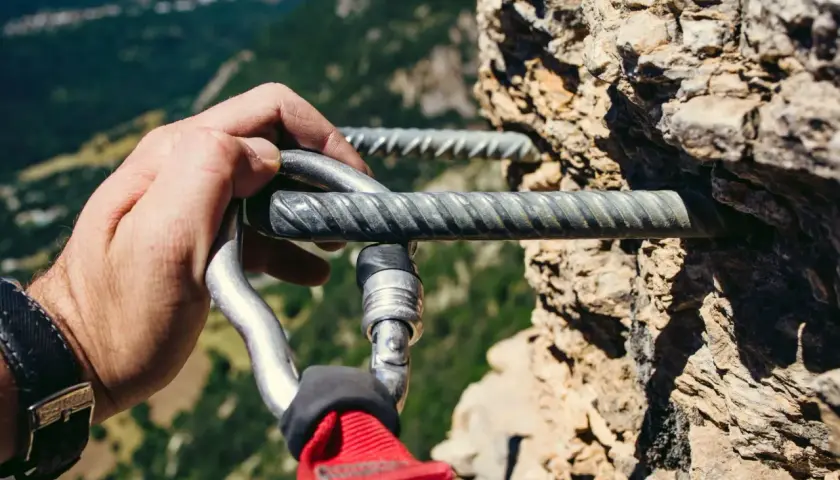 Mano enguantada se engancha a la **línea de vida vía ferrata** en una pared rocosa, con un paisaje montañoso al fondo.