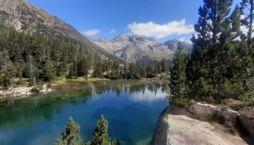 El ibonet de Batisielles, un espejo de agua turquesa rodeado de pinos y montañas imponentes, refleja el cielo azul y las nubes.