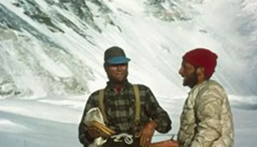 Dos montañistas sonríen en la nieve. Uno, con gorra y camisa de cuadros, sostiene un pico. El otro, con gorro rojo, parece ser Tom Hornbein.