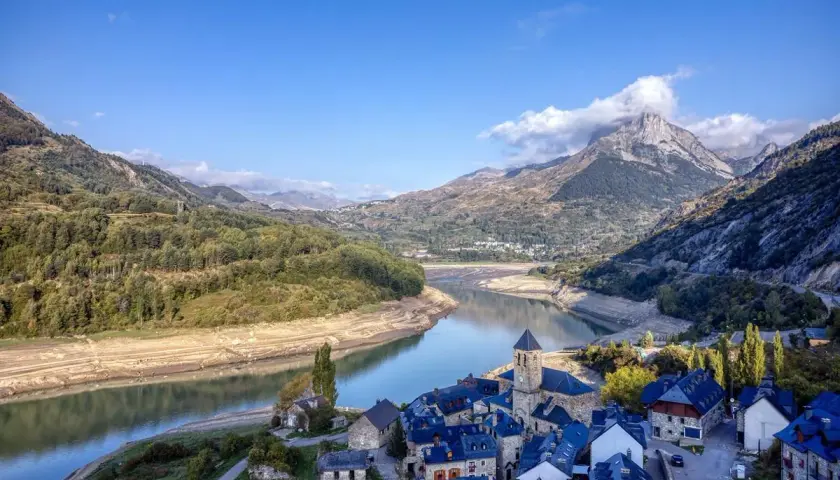 Pueblo de piedra junto a un embalse, con montañas nevadas de los picos de Huesca al fondo.