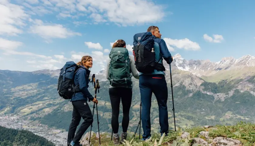 Tres excursionistas con mochilas y bastones admiran un paisaje montañoso. Sus prendas, ideales para distintos tipos de goretex, los protegen del clima.